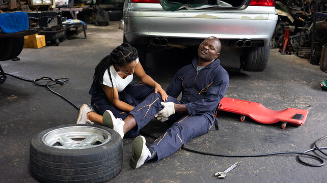 African American woman mechanic helped a black engineer man with knee pain due to work lie on the garage floor.