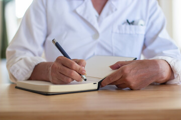 Fototapeta premium Close-up of professional female doctors hands during pandemic. Woman in medical uniform sitting at table with pen and notebook making notes. Doctors duty, health care, medical service concept