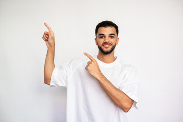 Portrait of positive young man pointing forefingers upwards. Bearded man wearing white T-shirt looking at camera and smiling over white background. Advertising concept