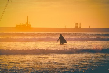 Silhouette of a surfer holding a surfboard standing in the wavy water at sunset