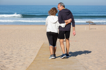 Back view of happy elderly couple resting on sea beach. Grey-haired man and woman walking together...