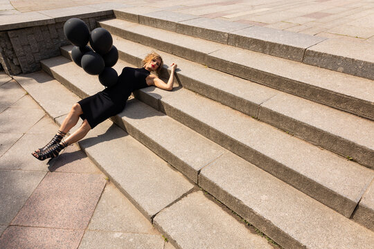 Beautiful woman in black dress with dark balloons lies on stairs in park. Art shooting from unusual angles. High quality photo