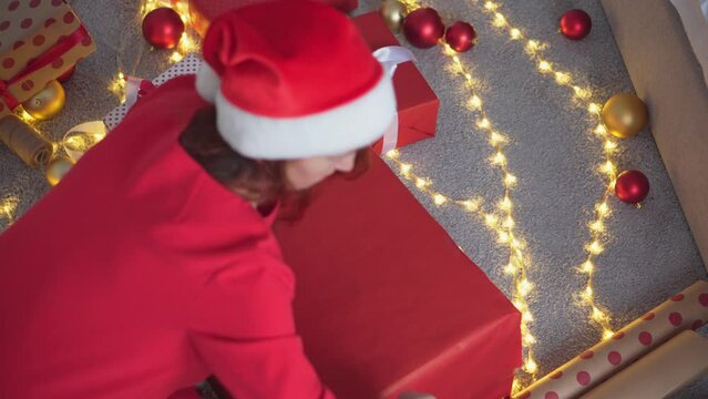 Girl in a Santa hat creates New Year's packages from red wrapping paper. Young woman is packing Christmas presents for her friends and relatives. Decorations, preparation for the winter holiday season