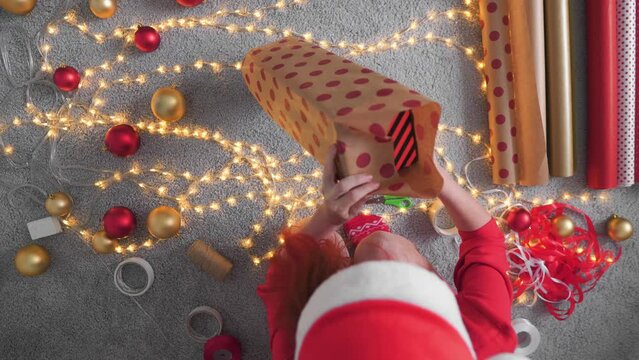 Girl in a Santa hat creates New Year's packages from red wrapping paper. Young woman is packing Christmas presents for her friends and relatives. Decorations, preparation for the winter holiday season