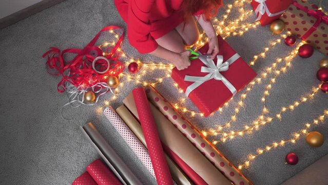 Girl in a Santa hat creates New Year's packages from red wrapping paper. Young woman is packing Christmas presents for her friends and relatives. Decorations, preparation for the winter holiday season