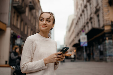 Fototapeta premium Young stylish woman in sweater using a smart phone standing outdoors on the street in Warsaw. Portrait of beautiful woman reading message on the street.