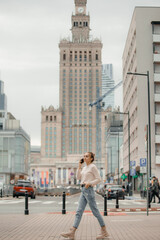 Obraz premium Young woman using mobile phone walking on the city street in Warsaw, Poland. Successful woman calling with cell telephone outside,business talking via mobile phone and walking on the city street.