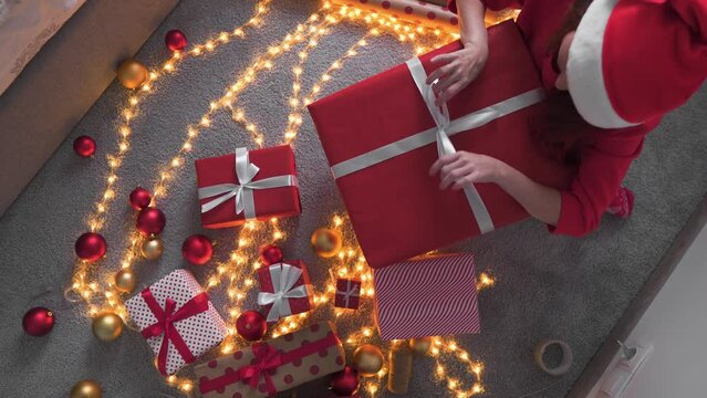 Girl in a Santa hat creates New Year's packages from red wrapping paper. Young woman is packing Christmas presents for her friends and relatives. Decorations, preparation for the winter holiday season