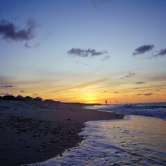 Shallow focus of a foamy wave crashing on a sandy beach, and a person walking, during the sunset