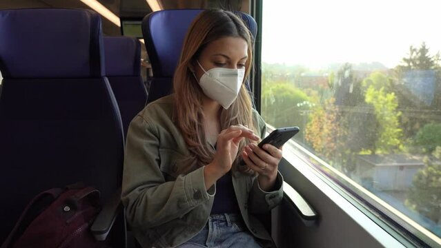Young woman with KN95 FFP2 face mask using smart phone app. Train passenger with protective mask traveling sitting in business class texting on mobile phone. Travel safely on public transport.