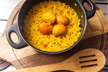 Uma panela preta com arroz com pequi e uma colher de madeira, sobre uma tábua de cozinha. Um prato típico brasileiro.