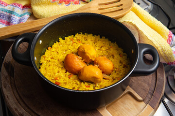 Uma panela preta com arroz com pequi, um pano de prato e uma colher de madeira, sobre uma tábua de madeira. Um prato típico brasileiro.