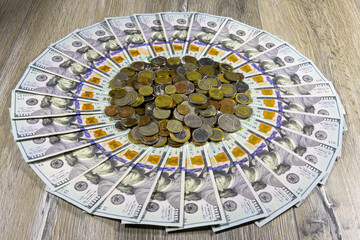 One hundred dollar bills of American dollars laid out in a circle with a bunch of different coins from around the world in the middle on a gray wooden