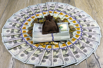 A money sack and a stack of American dollar bills on a pile of different coins from around the world. One hundred dollar bills laid out in a circle on a gray wooden table