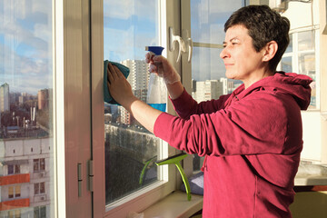 Middle aged brunette woman in rubber gloves washes window on the balcony in her apartment with rag....