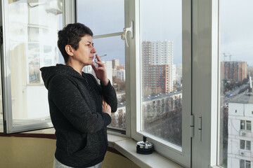 Middle-aged brunette woman stands on the glazed balcony of her apartment and smokes. A woman with a...