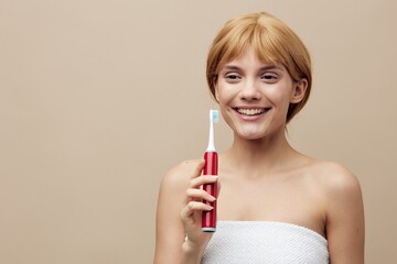 a joyful blonde woman with her hair pinned back is standing on a beige background wrapped in a towel and holding a red electric toothbrush in her hand, smiling and squinting her eyes with delight