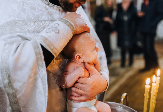 A male Christian priest in a church conducts a sacred rite, a ritual for a beautiful newborn, holding in his arms and lowering into a sacred font with water. Photography, religion.
