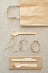 Topic of eco friendly, disposable, recycling, takeaway, plastic free. Craft paper bag, wooden spoon, knife, fork, paper napkin, word eco written with twine on canvas tablecloth. Top view.