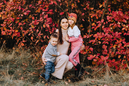 Photography, a portrait of a beautiful brunette girl with a small child, a boy, a son, and a smiling girl, a daughter, in nature in autumn, against the backdrop of a tree with red leaves.