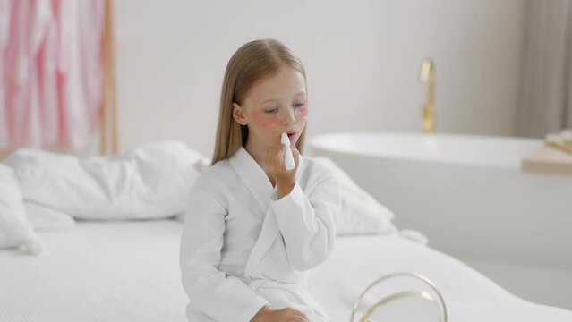 Adorable girl with patches under eyes applying lipstick on lips and sending blow kiss to reflection, empty space