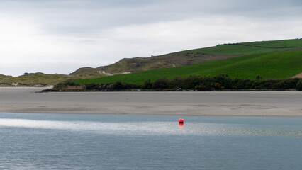 Fototapeta na wymiar A orange buoy on the shallows. Seaside landscape. Cloudy sky over a green hill. Ocean ebb.
