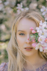 Fototapeta premium Portrait of a beautiful girl looking directly at the camera, blooming apple orchard, long blond hair, spring time