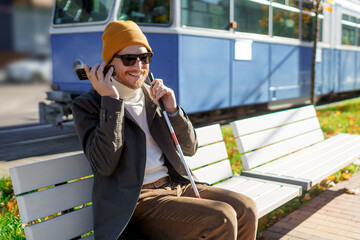 Blind man with a walking stick sits on a bench uses a smartphone on the background of a city tram