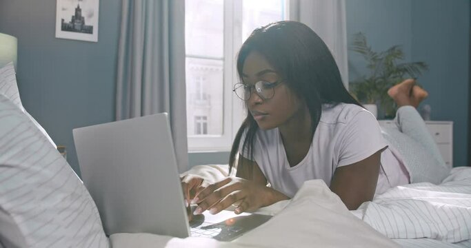 Portrait of successful pretty focused African woman working online on laptop lying in bed. Smart concentrated African-American female girl wearing glasses typing freelancing from home doing tasks.