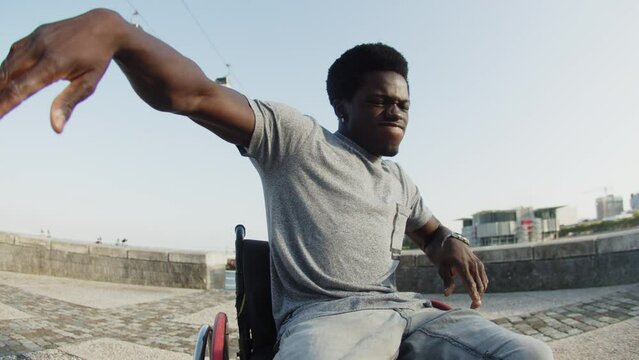 Happy African American man dancing using wheelchair outdoors. Young man with disability enjoying music at quay. Man with disability concept