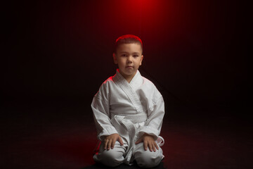 A little karate boy in a white kimono sits on his knees on a black isolated background.