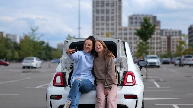 Cheerful Caucasian adult beautiful woman with teen girl kid looking at smartphone camera taking selfie photos sitting in car trunk. Mother and daughter posing to cellphone camera.