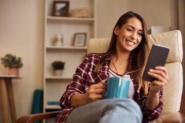 Happy woman texting on cell phone while having cup of coffee at home.
