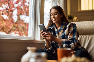 Smiling woman text messaging on smart phone while relaxing by window at home.
