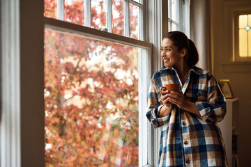 Happy woman looking through window while drinking tea at home.