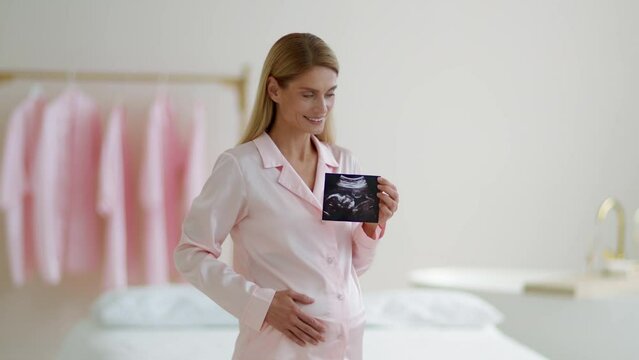 Middle aged pregnant woman in pajamas enjoying first photo of baby, holding sonography and caressing belly, free space