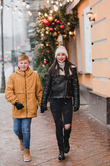 Caucasian mother and son in warm cozy clothes walk outdoors on festive city streets. Family walks at the fair as snow falls. Cold happy winter day. Holidays, Christmas, New Year, love concept.