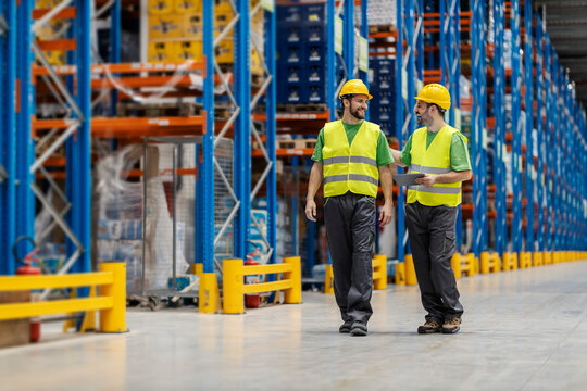 Two happy warehouse workers walk in facility with tablet and catting. Teamwork.