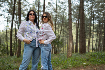 Naklejka premium Two model girls who wears same clothes are in forest . White shirt, jean and white boots. One of the is blonde, one of them has black hair. Model shoot. Fashion concept.