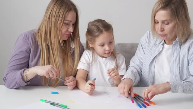 Family art therapy. Female generation. Enjoying time. Creative girl drawing with mother and grandmother with colorful markers sitting desk light room interior.
