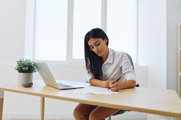 A woman works and learns by writing down text on paper with a pen and checking for errors, studying and teaching in college, business work at a desk with a laptop