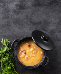 Pea soup with smoked ham in black soucepan on dark background with herb.