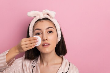 portrait beautiful young woman in pink pajamas on the background in the studio removes makeup from the face with a cotton pad.