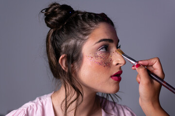 Portrait of brunette girl in makeup studio, makeup artist lays the foundation on skin with brush against background of bokeh light bulbs on mirror. Makeup and beauty. Lifestyle - obrazy, fototapety, plakaty