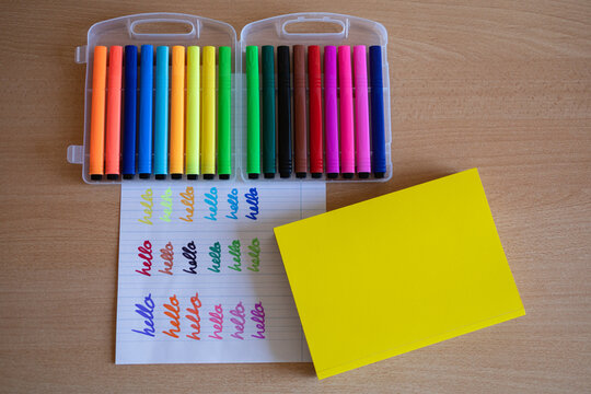 Box Of Colored Ink Markers Next To The Word Hello Written In Each Color On A Piece Of Paper. There Is A Yellow Cover Book With Copy Space For Text.