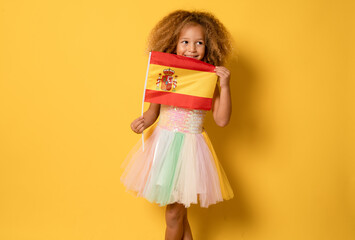 Cute little girl holding Spanish flag looking side standing isolated over yellow background.