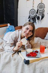 A young slender woman is sitting on the bed and eating a delicious breakfast of rice cakes and...