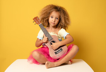Fototapeta premium Cute little girl in summer clothing playing ukulele over yellow background. Happy vacation concept.