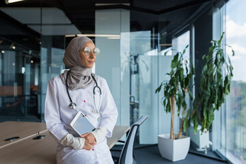 A happy and thoughtful young Muslim woman, an Arab doctor in a hospital, is sitting at her desk,...