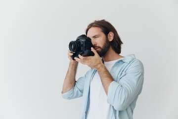 A male hipster photographer in a studio against a white background looks through the camera viewfinder and shoots shots with natural light from the window. Lifestyle work as a freelance photographer